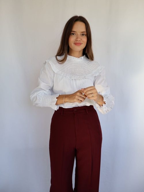 Mujer con blusa blanca de manga larga abullonada, cuello fruncido con volante y puños fruncidos, combinada con pantalón burdeos.